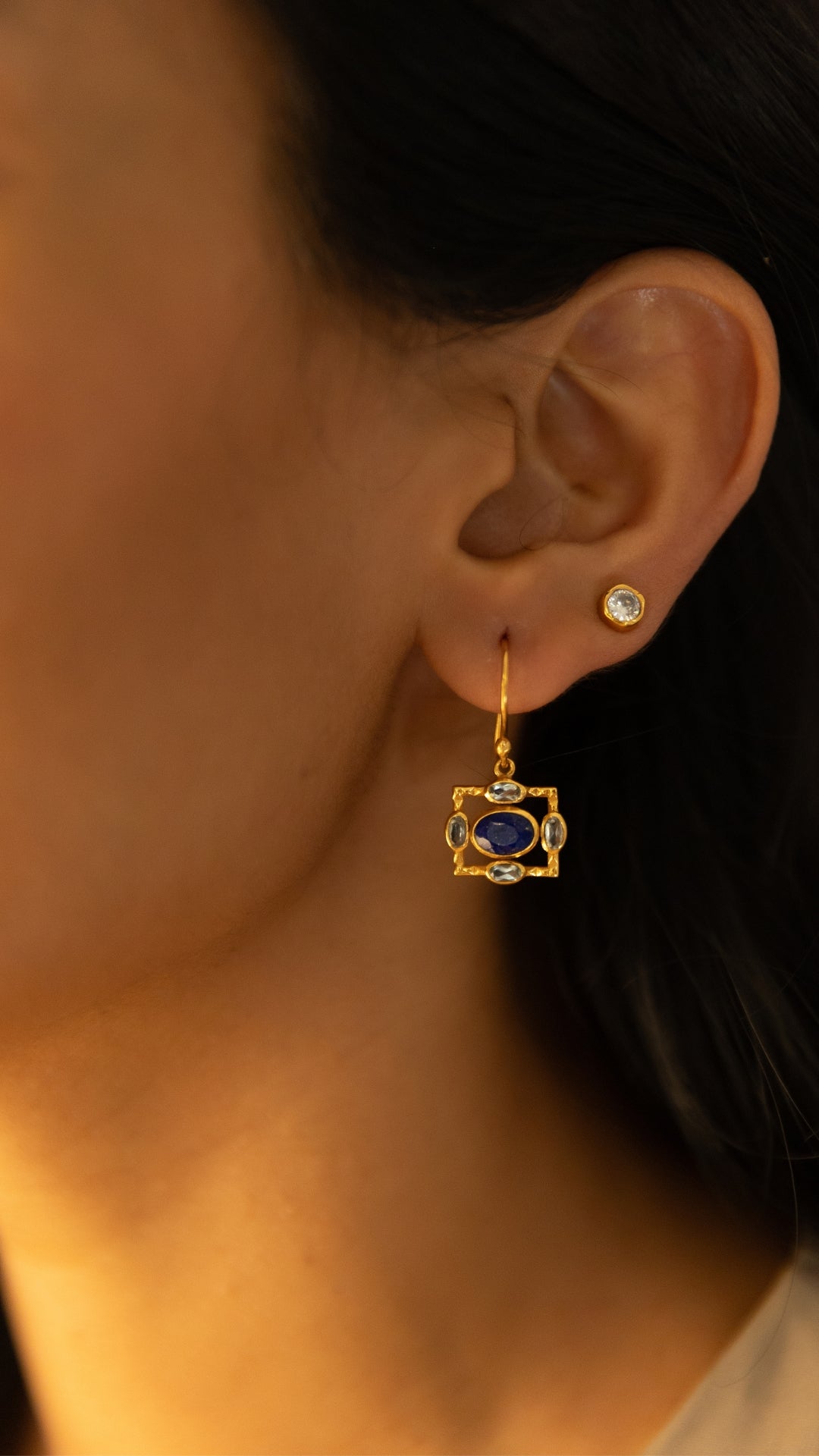 A close-up of a person's ear wearing a gold dangle earring with a blue oval center stone and small clear accent stones, alongside a simple gold stud earring.